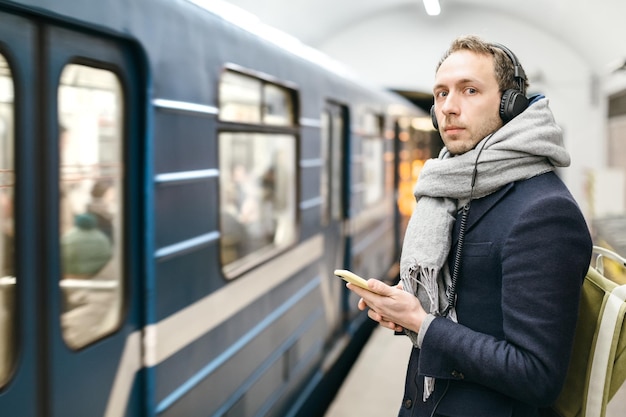Man die op het metroplatform staat en zijn smartphone in handen houdt, luistert naar muziek met een koptelefoon