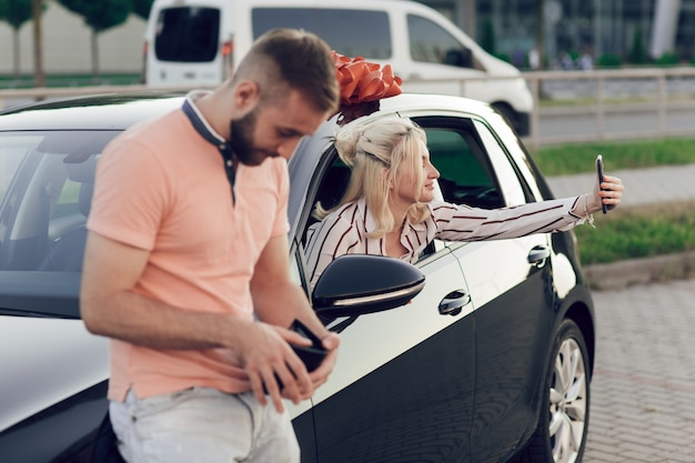 Foto man die vrouw verrast door een nieuwe auto te kopen. gelukkig jongedame achter het stuur zitten en het nemen van een selfie.