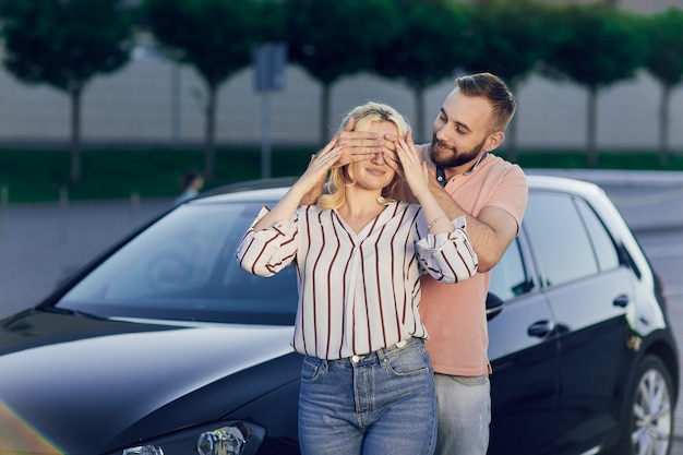 Man die vrouw verrast door een nieuwe auto te kopen. Jong stel koopt auto, man en vrouw staan naast de auto op straat. Verrassingsgeschenk voor een geliefde.