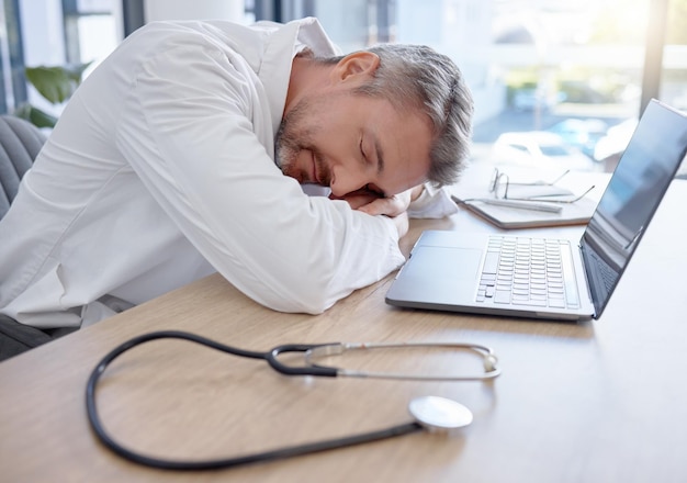 Photo man doctor and laptop sleeping on desk from burnout overworked or insomnia at the office tired male medical professional taking a nap rest or asleep on table by computer at the workplace