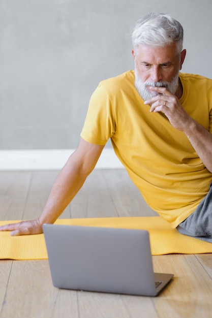 A man does yoga and fitness in the gym with a trainer online from a laptop senior male sports coach