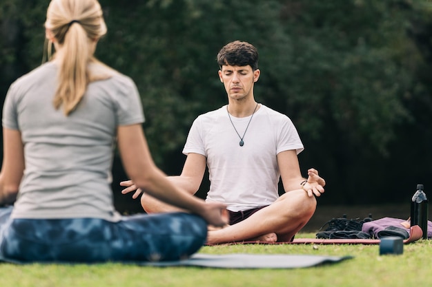 Man doet lotus yoga pose op een mat voor een vrouwelijke instructeur in een park omringd door bomen