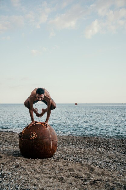 Man doet yoga oefening buitenshuis staande op oude roestige drijvende zeemijn op het strand met rocky