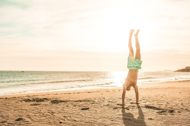 Man doing a headstand on the sand of the beach at sunset alone - healthy and nice lifestyle at summer