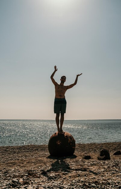 Man doing yoga exercise outdoors standing on old rusty floating\
marine mine on the beach with rocky