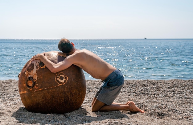 Man doing yoga and meditation outdoors near to old rusty floating marine mine on the beach with rocky shore and sea background Healthy lifestyle pollution nature protection war and peace concept