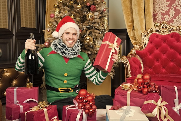 Man draagt kerstmuts elf kleren vieren kerstmis interieur versierde dennenboom en geschenken Kerst champagne drinken Man viert nieuwjaar of kerstvakantie Santa en kerstcadeaus concept