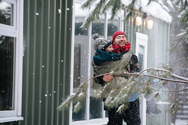 Man draagt zijn vrouw in handen. ze krijgen overal sneeuw van een nabijgelegen boom. Beiden dragen hoeden, jassen en sjaals. Voor een huis met golfplaten muur.