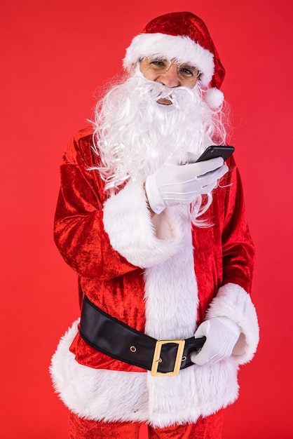 Man dressed as Santa Claus consulting his mobile phone on red background Christmas celebration gifts consumerism and happiness concept