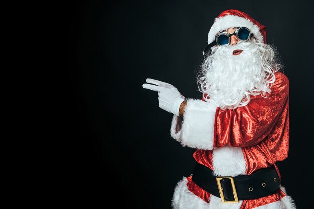 Man dressed as Santa Claus wearing Victorian style welding goggles, pointing to the left with his fingers, on black background. Christmas concept, Santa Claus, gifts, celebration.