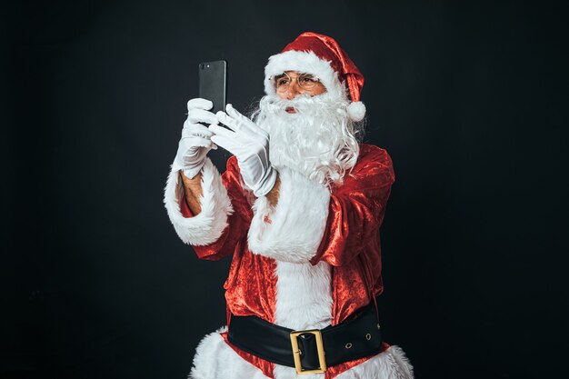 Man dressed as Santa Claus with a mobile in his hands, taking a selfie, on black background. Christmas concept, Santa Claus, gifts, celebration.