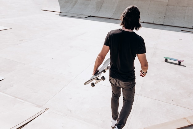 スケートボードを手にカジュアルな服を着た男が、外の晴れた日にスケートパークの滑り台を歩いている。