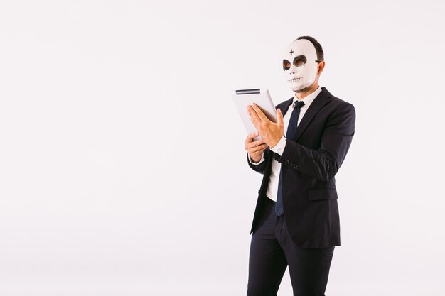 Man dressed in suit and tie, wearing a killer's mask with a cross on his forehead for Halloween, looking at his tablet. Carnival and halloween celebration