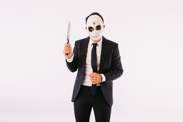 Man dressed in suit and tie, wearing a killer's mask with a cross on his forehead for Halloween, threatening with a knife. Carnival and halloween celebration