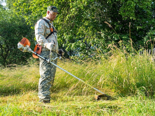 A man dressed in work clothes using a gasoline mower cuts long grass. Summer. rural area, maintenance of the territory