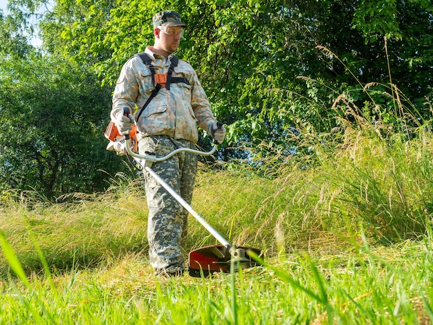 ガソリン芝刈り機を使用して作業服を着た男性が長い草を刈る夏の農村地域の領土の維持