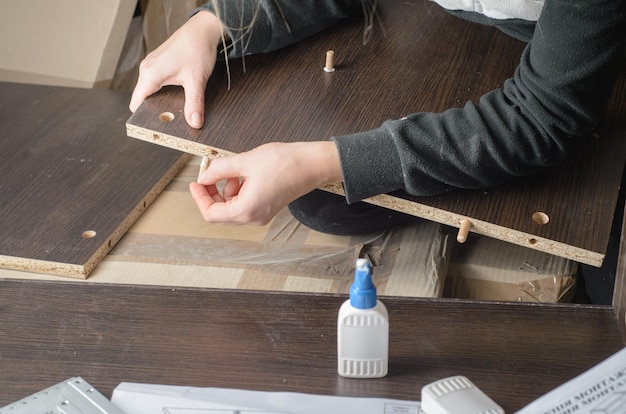 Photo man dressed in workers' overall assembling furniture sitting on the floor and glues furniture details