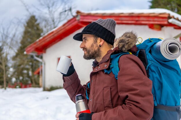 Man drinking coffee from a hot thermos in winter in the snow next to a cabin after sleeping in it