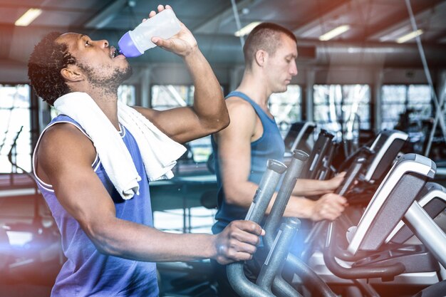 Man drinking water while using elliptical machine at gym