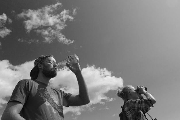 Foto uomo che beve acqua mentre donna guarda attraverso il binocolo contro il cielo.