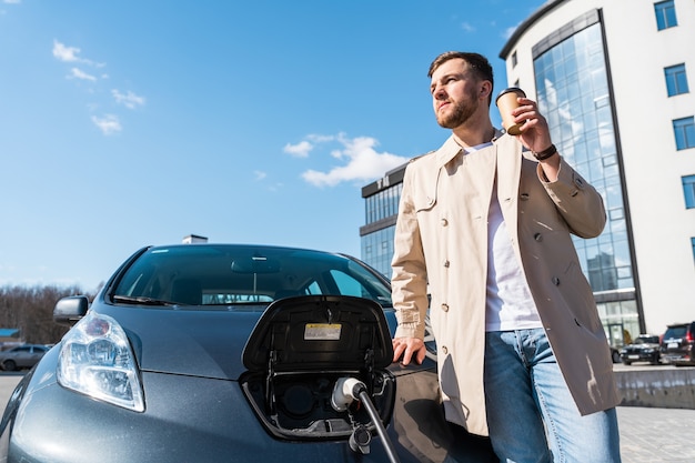 Foto l'uomo beve caffè mentre carica la sua auto elettrica