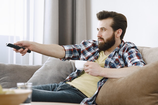 Man drinkt koffie. Guy tv kijken op de bank. TV-afstandsbediening in handen.