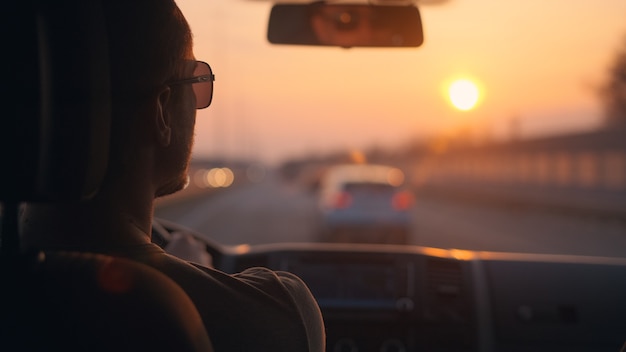 Мужчина водит машину по шоссе на фоне закатного неба