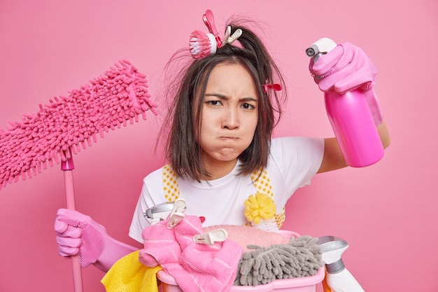 man drukt op zijn lippen ziet er moe uit terwijl hij de lente schoonmaakt houdt dispenser fles mop reinigt kamer poses in de buurt van wasmand geïsoleerd op roze