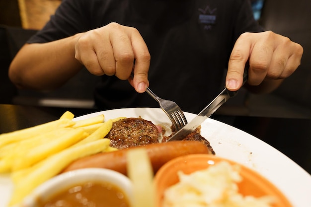 Мужчина ест мясо на гриле из тарелки, держа в руке нож и вилку, разрезая стейк из говядины на гриле