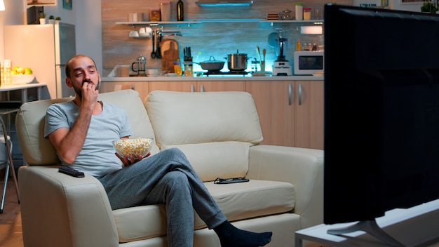 Man eating popcorn and watching TV, enjoying mid-night snack. Relaxed man lying on sofa late at night while watching entertainment tv shows at home cinema eating popcorn