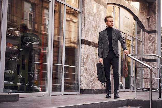 Man in elegant formal wear and with bag is outside against modern building.