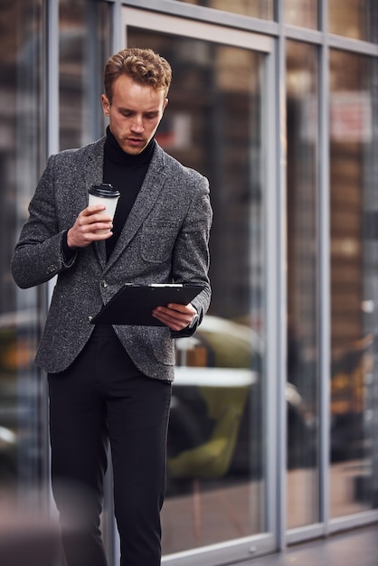 Man in elegant formal wear with cup of drink and notepad in hands is outside against modern building.