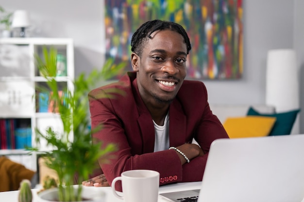 Man elegantly dressed in jacket sits at desk with a laptop and cup of coffee in the room