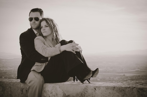 Photo man embracing woman while sitting on retaining wall against sky