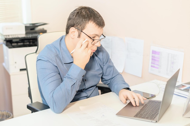 Man employee in blue shirt and glasses sitting at the desk in office using laptop computer Business busy working day concept