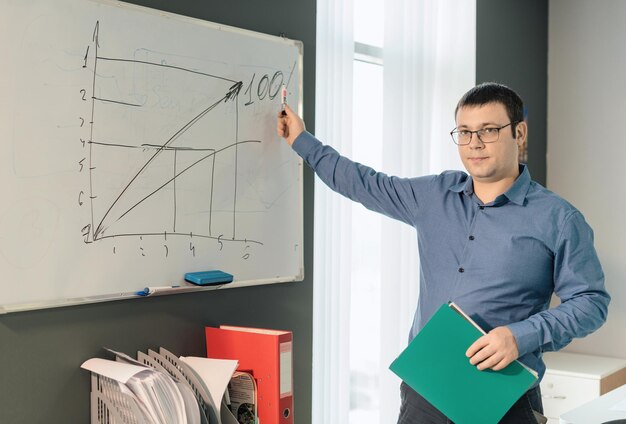 Man employee in blue shirt and glasses standing near the white\
board and points to the graph during business meeting in the office\
business busy working day concept