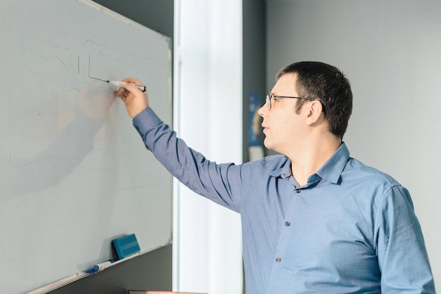 Man employee in blue shirt and glasses writing something on\
white board during business meeting in the office business busy\
working day concept