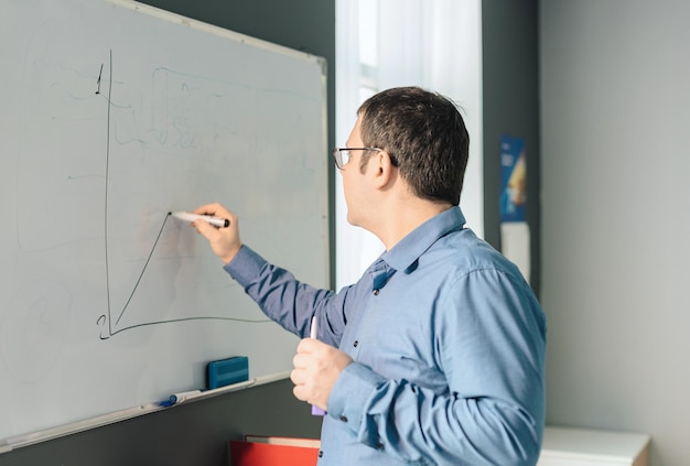 Man employee in blue shirt and glasses writing something on\
white board during business meeting in the office business busy\
working day concept