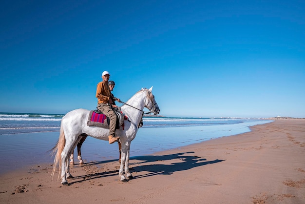 Man en vrouw die paarden berijden langs kustlijn bij strand tegen duidelijke hemel