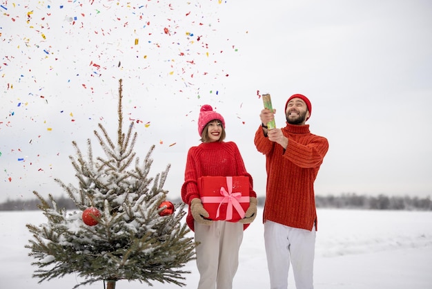 Man en vrouw hebben samen plezier door confetti te schieten bij de kerstboom in de buitenlucht. Jong echtpaar viert samen de nieuwjaarsvakantie.