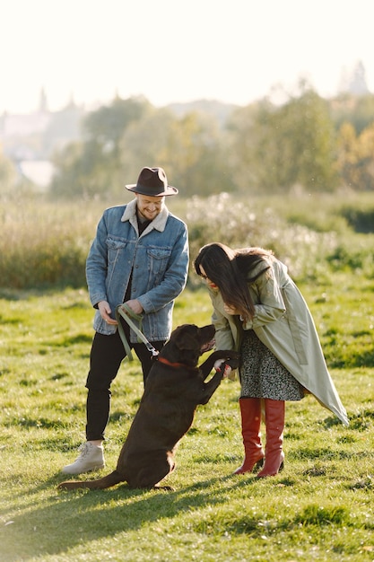 Man en vrouw in modekleren rustend op een natuur met hun labrador. Man met jas en zwarte hoed en vrouwenrok en rode panty