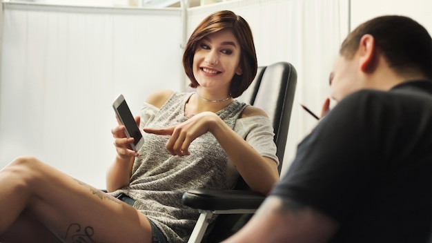 Man en vrouw kiezen tattoo ontwerp op telefoon in studio. Jong meisje toont foto op smartphone aan professionele mannelijke tatoeëerder, bespreekt schets van toekomstige bodyart, kopieer ruimte