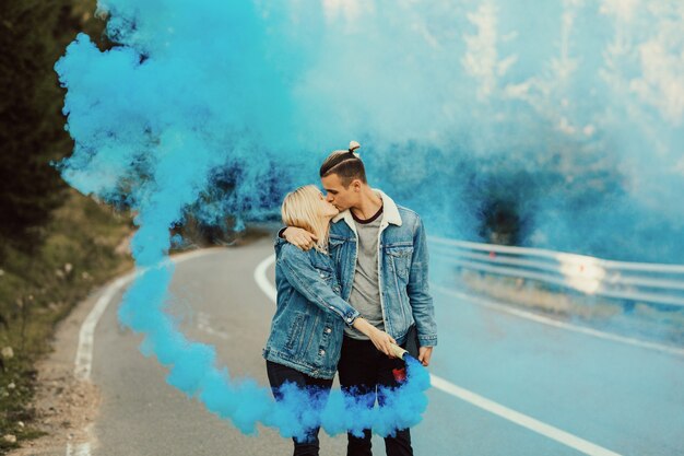 Man en vrouw kussen op de weg in de bergen met een blauwe kleurrijke rook in handen.