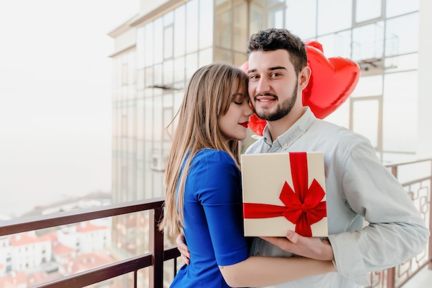 Man en vrouw met cadeau en rode hartvormige ballonnen op balkon thuis