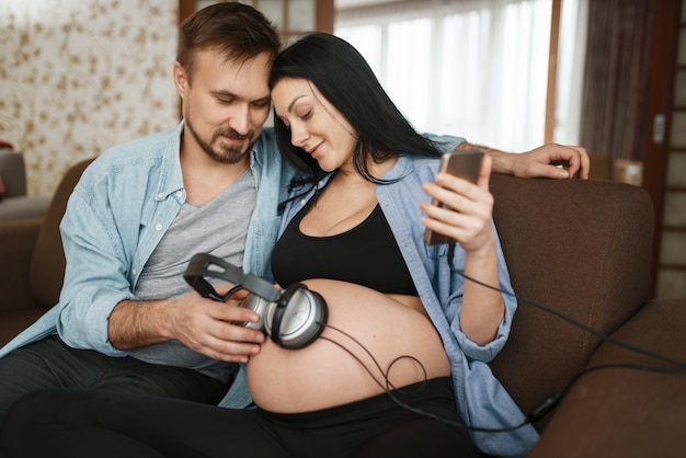Man en zwangere vrouw met koptelefoon op haar buik. Zwangerschap, prenatale periode thuis. Aanstaande mama en papa rusten op de bank, gezondheidszorg