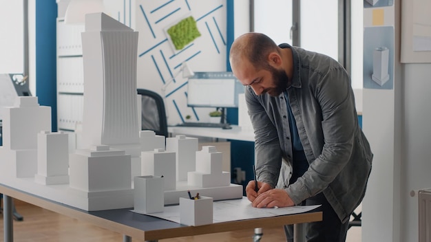 Man engineer analyzing building model to design blueprints plans on table. Architect working with maquette to plan construction layout and urban property development. Architecture project