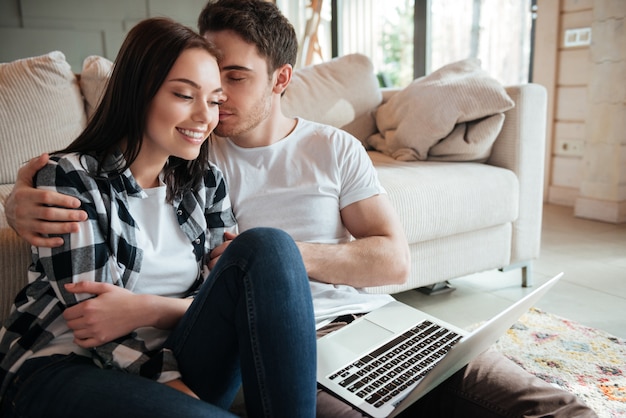 Man enjoying hugging his pretty woman with closed eyes on floor with laptop at home