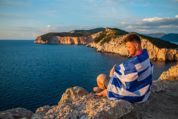 Мужчина наслаждается закатом над морем, сидя на скале, копируя космический флаг греции лефкас