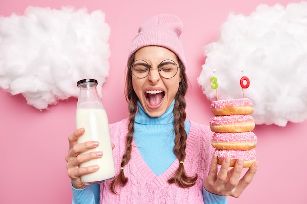 Photo man exclaims loudly celebrates birthday alone eats tasty donuts with milk keeps mouth widely opened wears hat vest and turtleneck round eyeglasses stands indoor