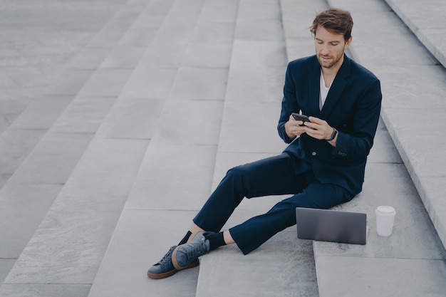 Man executive worker uses mobile phone for chatting works remotely makes project via laptop computer